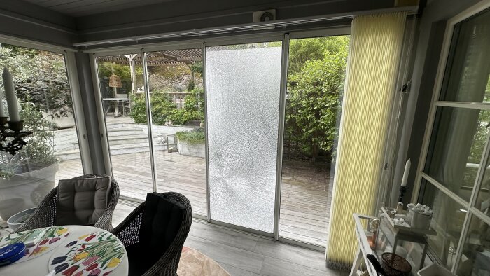 Sprucken glaspanel i skjutdörr med utsikt över terrass och trädgård. Inomhus syns stolar och dekorerat bord.