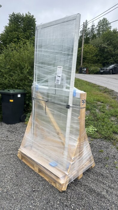 Inplastad glasad dörr levererad av Willab Garden, står på en träpall utomhus, redo för montering efter kundservicehantering.