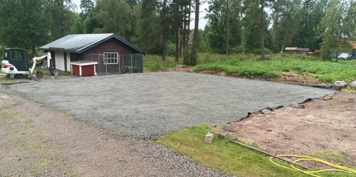 Nylagd grusplan framför ett mindre träskjul, omgiven av grönska och med en grävmaskin i bakgrunden.