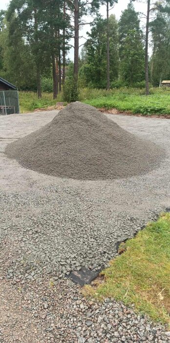 Hög med grus på en gård, omgiven av träd och grönt gräs. Gruset ska användas för ett byggprojekt.