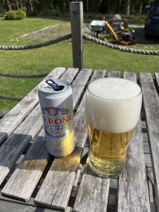Öppnad ölburk och glas med skummande öl på en träbänk utomhus, i bakgrunden syns en gräsmatta, skottkärra och trädgårdsredskap.