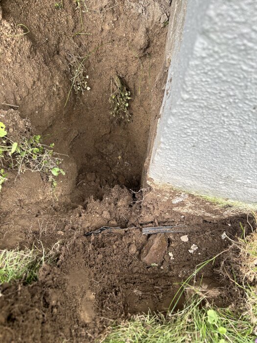 Grävd grop vid husgrund, exponerar jord och en bit av dräneringssystemet med brist på platonmatta och blottad husvägg.