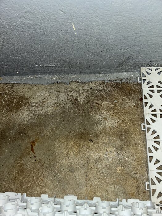 Betonggolv i källare med fuktfläckar och del av vit plastplatta synlig i hörnet.