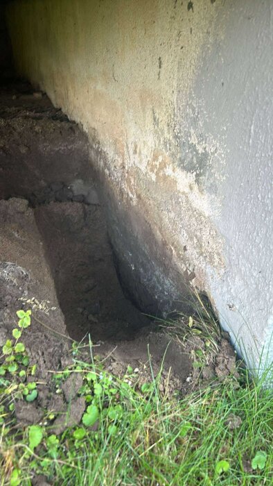 Grävd grop vid en husgrund med jordrester och gräs, synlig fukt på husväggen.