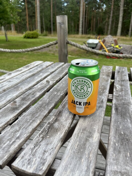 En burk Jack IPA-öl står på ett träbord utomhus med bakgrund av en trädgård och skogslandskap.