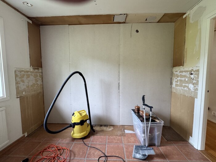 Vägg med nyligen installerade gipsskivor, en gul dammsugare, plastlåda med verktyg och sladdar på ett rödbrunt klinkergolv i renoverad rum.