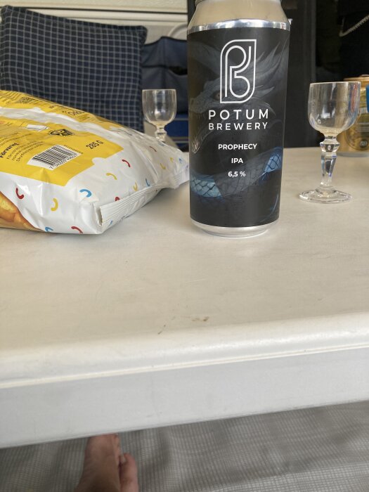 En ölburk från Potum Brewery står på ett bord bredvid ett chipspaket och två tomma glas.