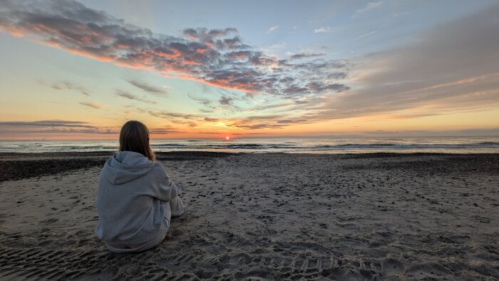 Person i grå huvtröja sitter på en sandstrand och tittar mot solnedgången vid havet med färgglada moln på himlen.