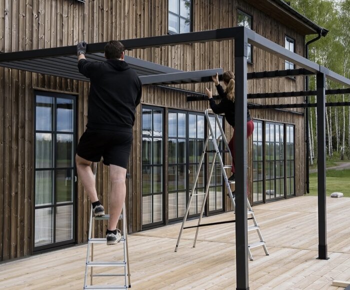 Två personer monterar en pergola på en träveranda vid ett hus med träfasad. De står på stegar och justerar takbalkarna.