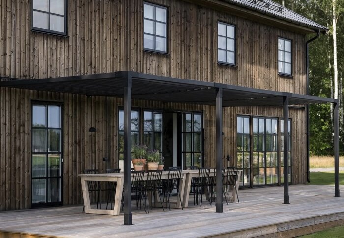 En svart pergola intill ett hus med träfasad, med svarta glasdörrar och fönster. Under pergolan står bord och stolar på en träaltan.