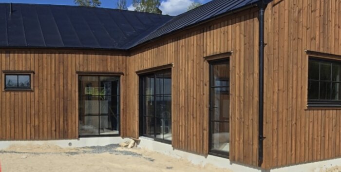 Trähus med vinkel och stora fönster mot trädgården, under diskussion för pergolanslutning vid fasad; sand och stenar ligger framför.