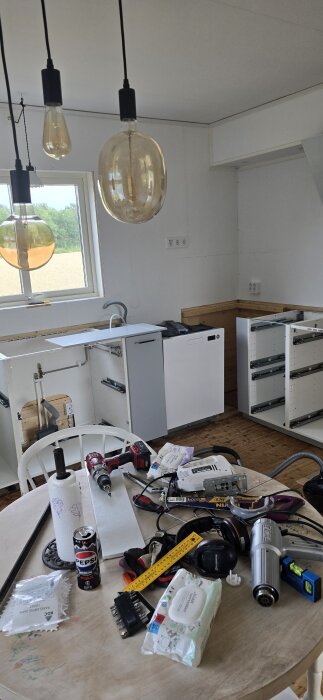 Verktyg och material på köksbord, omoderniserat kök med exponerade lådor, diskbänk, fönster och hängande lampor ovanför.