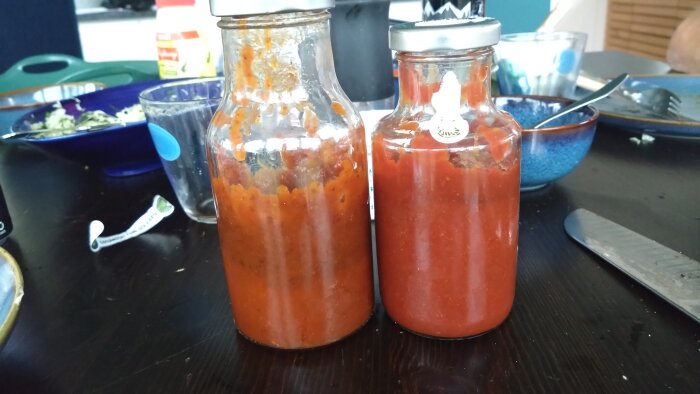 Två glasburkar med hemlagad röd bbq-sås på ett mörkt köksbord, omgivna av tallrikar och bestick.