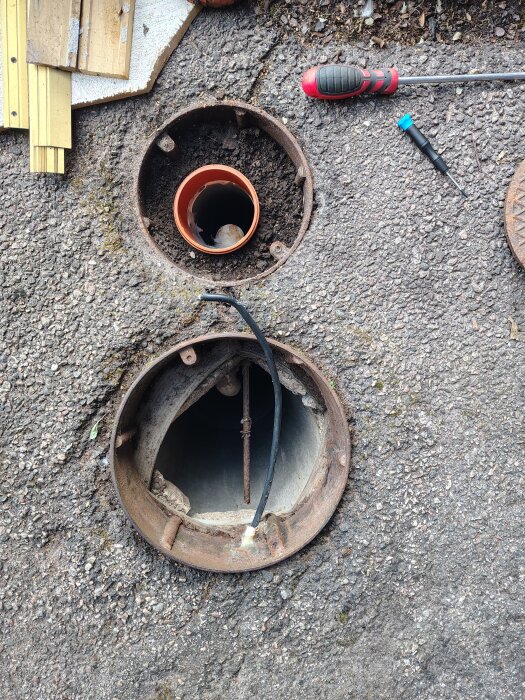 Öppet brunnslock med synliga rör, kablar och järnrör, omgivna av verktyg och byggmaterial på marken.