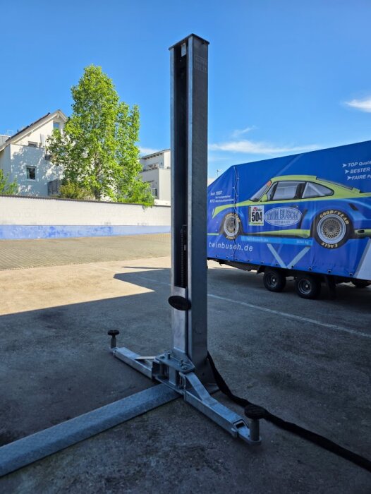En galvaniserad tvåpelarlyft utomhus på asfalterad yta framför en blå täckt släpvagn med motiv av en bil.