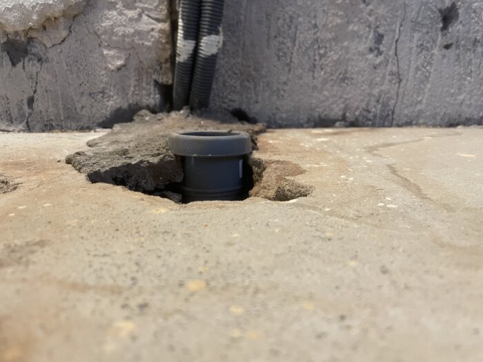 Avloppsrör i köksgolv nedanför betongyta med skarv synlig nära vägg, omlagd betong runt rör och eluttag/slingor i bakgrunden.