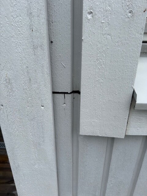 Spricka i träfasaden under en horisontell fönsterbräda på ett hus. Fasaden är vitmålad och sprickan är några centimeter bred.