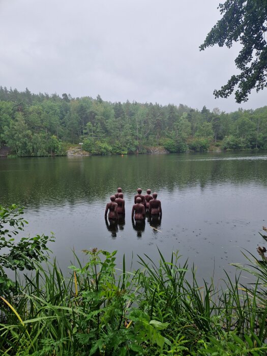 Skulpturer av människor står i vattnet vid en skogssjö, omgivna av grönska och träd, en mulen dag.