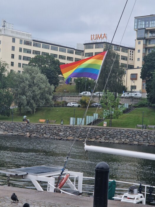 Regnbågsflagga vajar vid kajkant med vattendrag i förgrunden och byggnad med texten "LUMA" i bakgrunden.