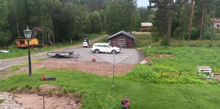 Grusplan med parkerad vit bil och släpvagn framför litet trähus, omgiven av träd och gräsmatta med en torkställning i förgrunden.