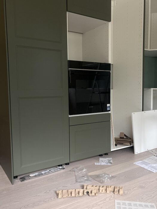 Grönt Ikea-kök med Siemens-ugn installerad, glipa ovan och nedan. Byggmaterial spritt på trägolv. Utrymme för mikrovågsugn synligt ovan.
