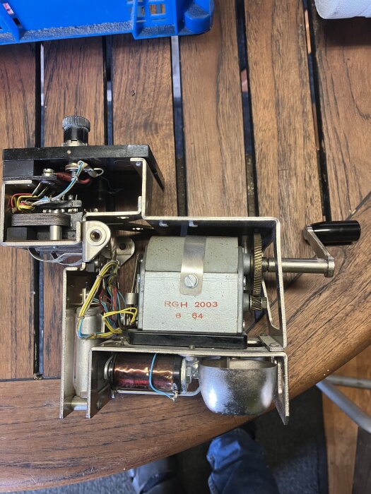 En mekanisk enhet med växlar, vev och ledningar, eventuellt en ringklocksmekanism, ligger på ett träbord.