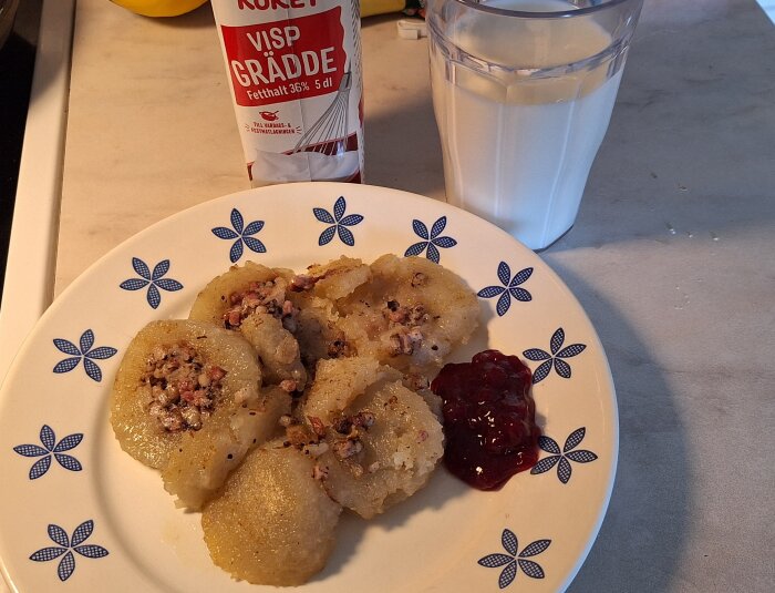Tallrik med kroppkakor, lingonsylt och grädde, bredvid ett glas mjölk.