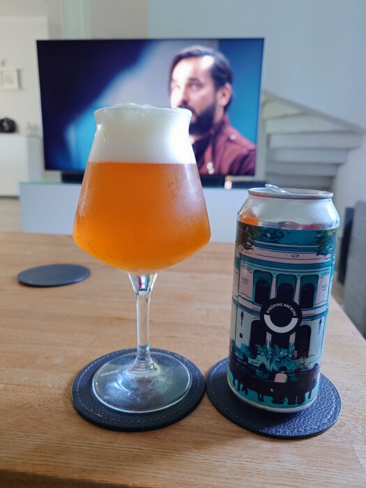 Ölglas med skummig DIPA från Nyköping Brewing, bredvid en dekorerad ölburk, står på ett bord framför en tv-skärm med en man i bakgrunden.