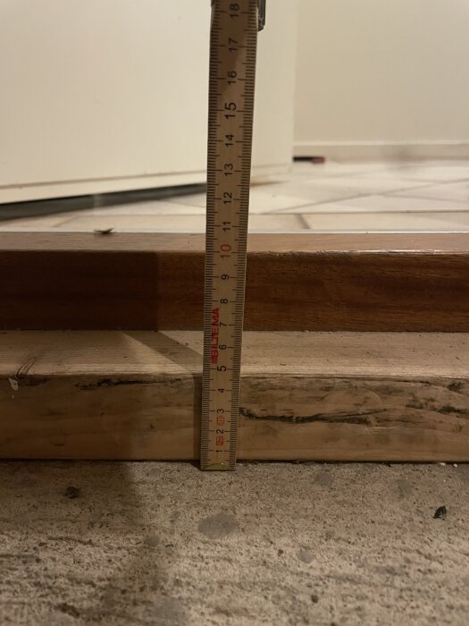 Måttstock mäts mot höjd på golvlist i trä, visar cirka 14 cm. Bakgrund med vitt kakelgolv och vit vägg.