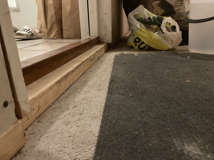 Dörrtröskel mellan två rum, golv med grå matta, plastpåsar med byggmaterial och ett par tofflor i bakgrunden.