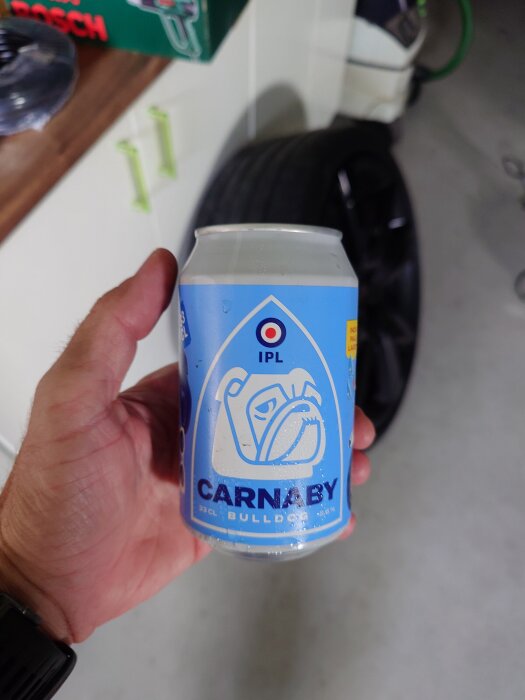 En hand håller en burk Carnaby Bulldog öl med blå etikett. I bakgrunden syns en del av ett garage med ett bildäck och verktyg.