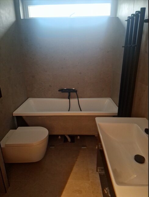 Ett badrum med modernt badkar, WC och handfat. Bakom badkaret en fönsterlucka, runtom ljusa kakelplattor.