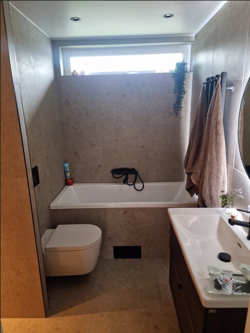 Ett modernt badrum med ett badkar, duschslang, hängande handdukar och ett fönster ovanför. Väggar och golv är i ljus sten.