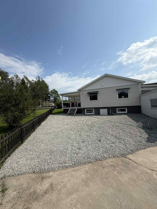 Uppfart med gårdsgrus framför ett vitt hus med veranda, omgiven av staket och gräsmatta. Klart väder med få moln på himlen.