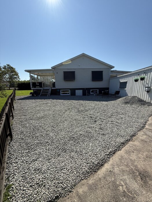 Grusad uppfart vid hus med vit fasad och altan. Solig dag, staket på sidan och trädgårdsmöbler syns.