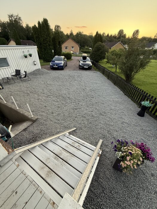 Grusad uppfart med två parkerade bilar, omgiven av grönska. Framför huset står blommor och en ramp som leder ner till gårdsplanen.