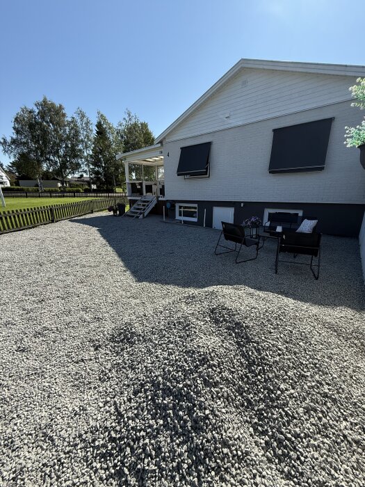 Hus med vit fasad, mark med grusad uppfart, sittgrupp, trappa och markiser. Träd och staket syns i bakgrunden mot klarblå himmel.