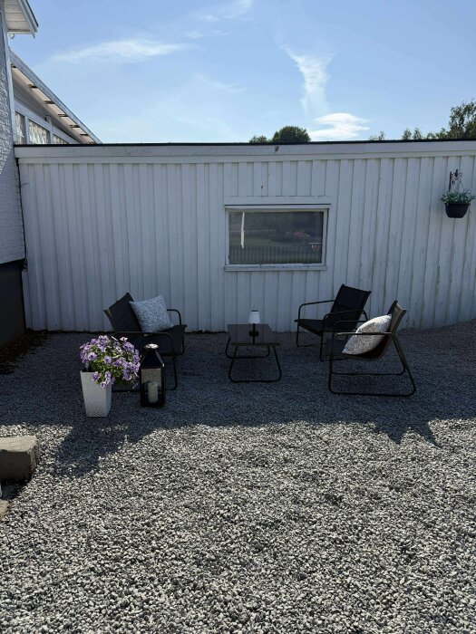 Trädgårdsmöbler på en grusad uteplats framför en vit byggnad, dekorerad med blomkrukor och en lykta.