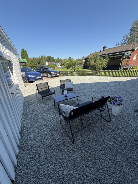 Grusad uppfart med utemöbler och blomkruka, två bilar parkerade i bakgrunden, solsken och hus samt trädgård runtom.