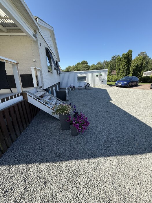 Grusad uppfart med blomkrukor kantar ett vitt hus. I bakgrunden syns träd, en bil och en sittgrupp mot ett vitmålat plank.