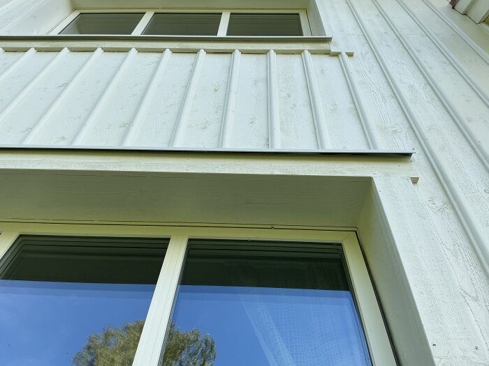 Vitmålad träfasad med fönster, nyligen målad med en strykning. Fönstret speglar blå himmel och träd.