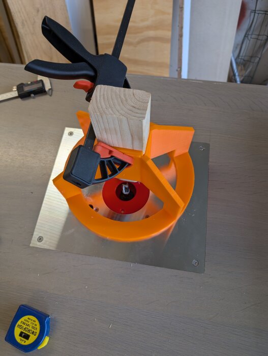 Träkloss klämd med svart och röd tving på orange 3D-utskrivet fäste, ovanpå metallplatta. Måttband och skjutmått på träbord.