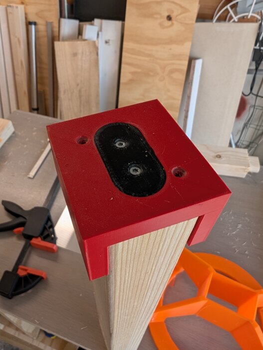 3D-utskrivet rött fäste ovanpå en träregel, del av projekt för fräsbord; omgivande verktyg och arbetsmaterial syns i bakgrunden.