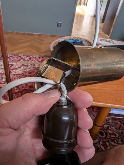 En hand håller upp en lamphållare med sladd och metallbeslag i bakgrunden av en matta och bord.