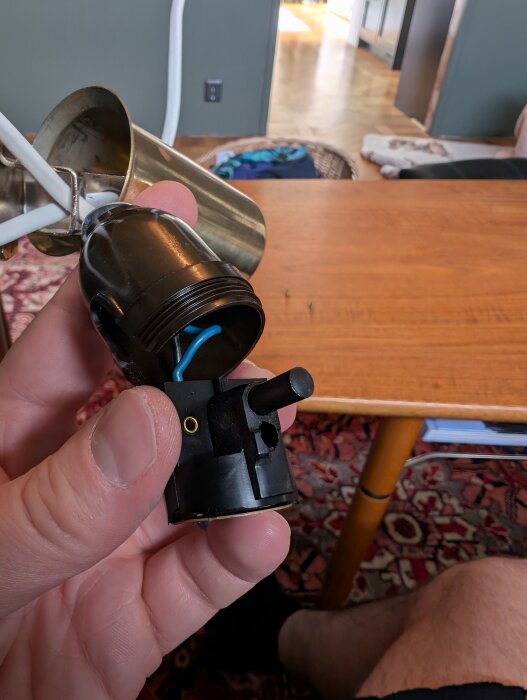 Svart lamphållare med rund tryckknapp och utvändig gänga visas i närbild mot en bakgrund av ett bord och golv.