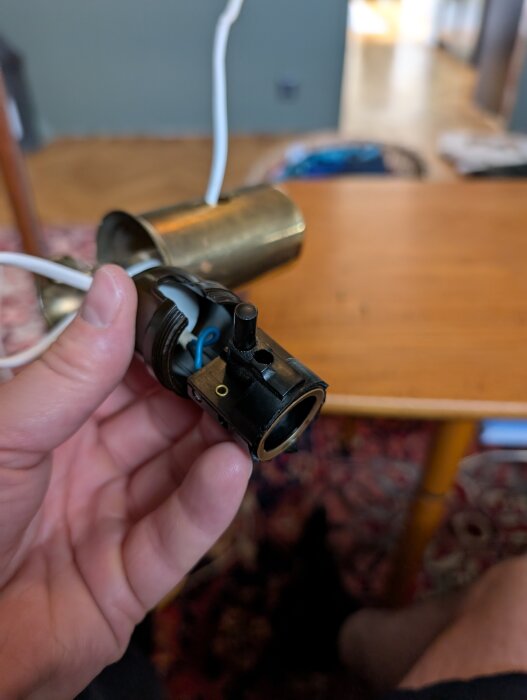 Hand håller en svart lamphållare med rundtryckknapp och sladdar, liggande på ett träbord inomhus.