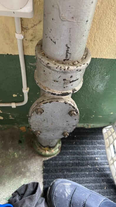 Gammalt avloppsrör i metall med rost och en renslucka fäst med bultar, placerat mot en gul och grön vägg.