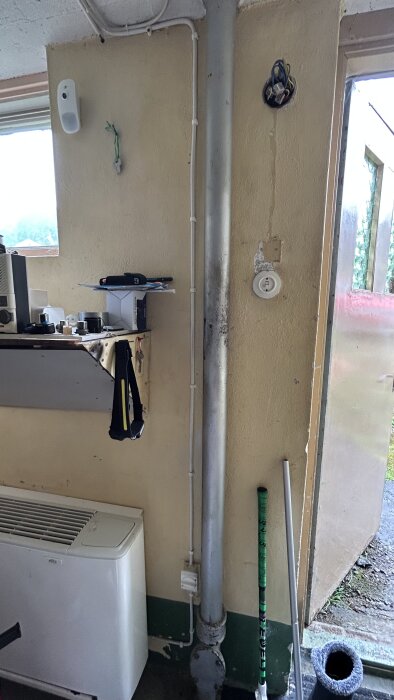 Metallrör går från golv till tak i ett rum med beige väggar. Hylla med verktyg och elektriska kablar syns också.