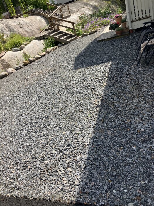 Gårdsplan med små stenar och en trätrappa i bakgrunden, omgiven av växter och gruslagt utrymme.