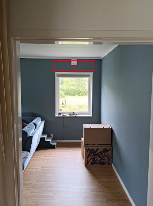 Rum med blå vägg och trägolv, ett fönster mitt på väggen, en soffa till vänster, och en kartong märkt "Biltema" till höger.